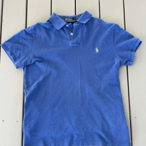 Blå pikétröja från Ralph Lauren - Säljer en klassisk blå pikétröja från Ralph Lauren med den ikoniska gröna loggan på bröstet. Tröjan har korta ärmar och en stilren krage. Perfekt för en avslappnad och stilren look. Går att diskutera om pris!