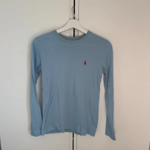 Ljusblå långärmad tröja från Ralph Lauren - Säljer en stilren ljusblå långärmad tröja från Ralph Lauren. Tröjan har en liten röd logotyp på bröstet och är perfekt för en avslappnad look. Materialet är mjukt och bekvämt, vilket gör den idealisk för vardagsbruk. Passar perfekt till jeans eller chinos.