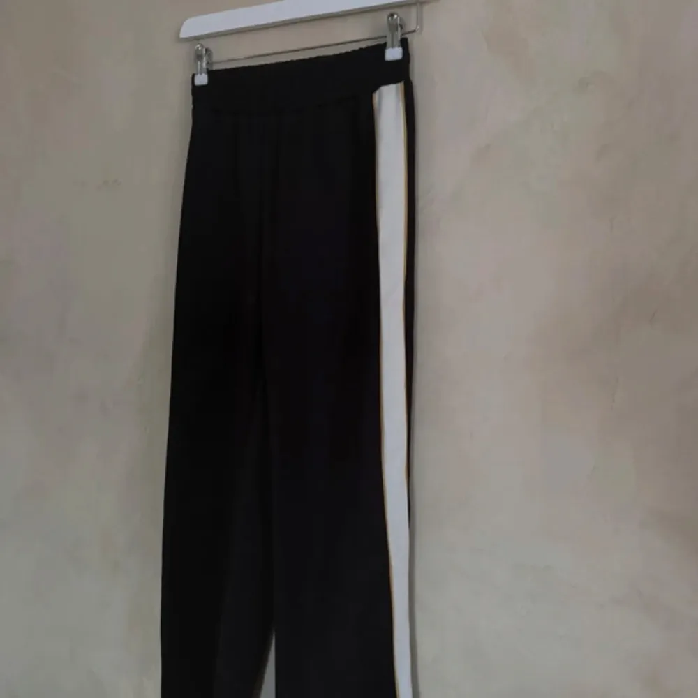 Snygga svarta byxor med vita ränder längs sidorna. De har en elastisk midja för extra komfort och en avslappnad passform. Perfekta för en sportig look eller en avslappnad dag.. Farkut & Housut.