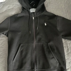 Svart hoodie från Polo Ralph Lauren - Säljer en klassisk svart hoodie från Polo Ralph Lauren med dragkedja och broderad logga på bröstet. Tröjan har en justerbar huva och praktiska fickor framtill. Perfekt för en stilren och bekväm look.