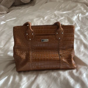 Brun handväska från Osprey - Snygg brun handväska från Osprey london med krokodilpräglat mönster. Väskan har två handtag och flera fack med dragkedja inuti för praktisk förvaring. Perfekt för att hålla ordning på dina saker med stil. Lite sliten vid handtagen men annars väldigt bra skick!💕