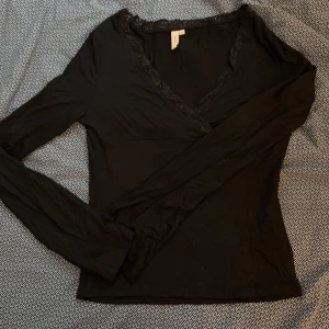 Svart långärmad topp med spetsdetaljer - Säljer en elegant svart långärmad topp med v-ringning och spetsdetaljer runt halsen. Perfekt för en stilren look. Passar bra till både jeans och kjol. Aldrig använd!