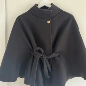 Svart kappa från Missay - Elegant svart kappa från Missay med hög krage och knäppning med en dekorativ knapp. Kappan har ett knytbälte i midjan för en justerbar passform.