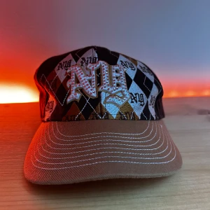 NY keps brun - Snygg brun keps med rutigt mönster i svart, vitt och brunt. Kepsen har en broderad 'NY' text framtill och en mindre broderad text baktill. Tillverkad i bomull och perfekt för en cool och avslappnad stil.