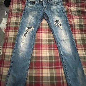 Blå jeans från Mishumo - Snygga blå jeans från Mishumo med slitna detaljer och en klassisk femficksdesign. Tillverkade i 100% bomull för en bekväm passform. Perfekta för en avslappnad stil med en touch av edge.