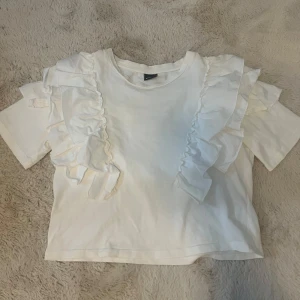 Vit topp med volangdetaljer från Gina Tricot - Säljer en söt vit topp från Gina Tricot med volangdetaljer längs axlarna. Toppen har korta ärmar och en bekväm passform. Perfekt nu till våren och sommaren💗