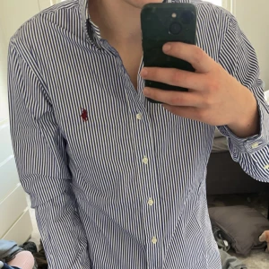 Blå och vit randig skjorta från Ralph Lauren - Säljer en stilren blå och vit randig skjorta från Ralph Lauren. Skjortan har en broderad logotyp på bröstet och är tillverkad med slim fit passform. Perfekt för en klassisk look.