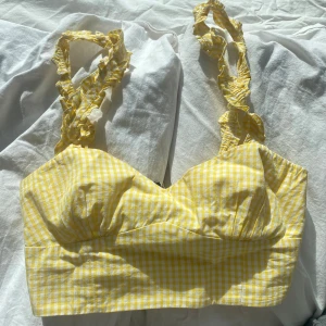 Gul rutig topp med volangdetaljer - Säljer en söt gul rutig topp med volangprydda axelband.. Den har en smickrande skärning och elastisk rygg för extra komfort. 