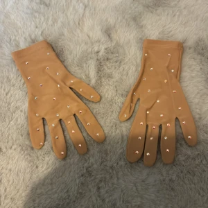 Beige handskar med strass från Sagester - Hudfärgade tävlingshandskar applicerade med äkta AB Swarovskikristaller. Kristallerna medför att hanskarna blir väldigt effektfulla samtidigt som händerna slipper vara bara. Finns ett litet hål i almen på ena handsen men utöver det är dom i mycket gott skick. Nypris 599