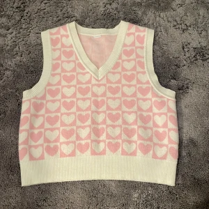 Rosa och vit stickad väst med hjärtmönster - Söt stickad väst i rosa och vitt med ett hjärtmönster. Västen har en v-ringad design och ribbade kanter. Endast använd ett fåtal gånger. Storlek 146-152