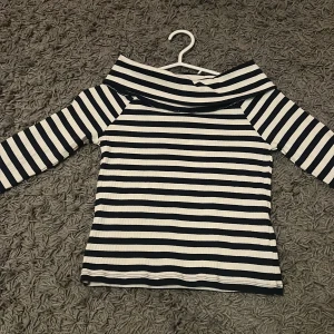 Randig offshoulder topp - Snygg randig topp med offshoulder-design i svart och vit. Perfekt för en stilren look med trekvartsärmar och ribbad textur. Passar bra till både jeans och kjol för en avslappnad men chic stil. Den är också oanvänd. Med prislapp.