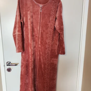 Mörkrosa morgonrock klänning från Romeo & Life - Säljer en elegant rostfärgad klänning från Romeo & Life med vackra broderade detaljer längs ena sidan och ärmsluten. Klänningen har en dragkedja framtill och är tillverkad i en mjuk bomulls- och polyesterblandning. Perfekt för en stilfull look!