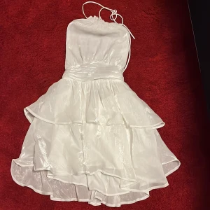 Vit axelbandslös klänning - Säljer en vit axelbandslös klänning i storlek 36/S med volanger och glansigt material💖✨🪩Klänningen har en kort längd och knytband vid halsen. Perfekt för sommarens festligheter såsom student, midsommar och liknande!🥳🎓