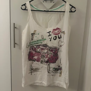 Vit linnetopp med tryck - Säljer en vit linnetopp med ett färgglatt tryck av en väska och texten 'I love you'. Toppen har en avslappnad passform och är perfekt för varma dagar.