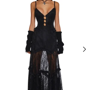 Svart spetsklänning med detaljer från dollskill - Elegant svart långklänning med spets och djup v-ringning. Klänningen har smala axelband och dekorativa knappar framtill. Den är delvis genomskinlig med ett vackert mönster och har en öppen rygg. Perfekt för en speciell kväll!