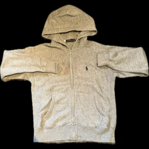 Grå hoodie från Polo Ralph Lauren - Säljer en stilren grå hoodie från Polo Ralph Lauren. Den har en dragkedja framtill och en klassisk logga på bröstet. Perfekt för en avslappnad look. 🐎