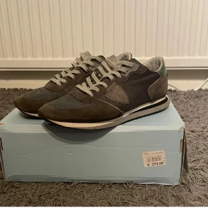 Bruna sneakers från Philippe Model - Snygga bruna sneakers från Philippe Model med grå snörning och detaljer i mocka. Skorna har en rund tå och en stilren design som passar perfekt för en avslappnad look. De har även en grön detalj vid hälen som ger en extra touch.