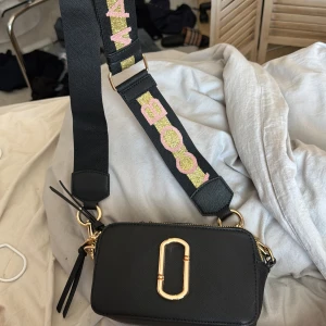 Svart axelväska från Marc Jacobs - Snygg svart axelväska från Marc Jacobs med en justerbar axelrem dekorerad med glittriga bokstäver i rosa och guld. Väskan har en praktisk dragkedja och en elegant guldfärgad detalj på framsidan. Perfekt för att bära dina nödvändigheter med stil.