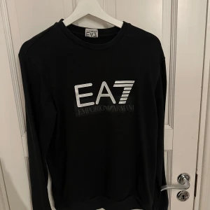 Svart långärmad tröja från Emporio Armani - Snygg svart långärmad tröja från Emporio Armani med stort EA7-tryck på framsidan. Perfekt för en stilren look. Passar bra till både jeans och chinos. 