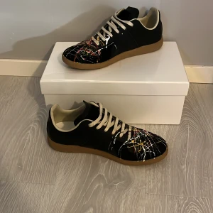 Svarta sneakers med färgstänk från Maison Margiela - Unika svarta sneakers från Maison Margiela med färgstänk i vitt, rött och gult. Skorna har en rund tå och snörning, vilket ger en cool och konstnärlig look. Perfekta för dig som vill sticka ut med stil. Dessa skick skulle jag säga är 8,5 eftersom märket på bild 3. Hör av med om ni har fler frågor. Priset är inte fast. Hör av er innan köp eftersom att jag bara kan frakta på vissa datum. 