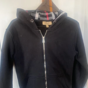 Burberry zip up hoodie svart - Snygg svart hoodie från Burberry med dragkedja och klassiskt rutigt mönster på insidan av huvan. Perfekt för en stilren look med en touch av lyx. Broderad logga på bröstet. Passar mer som xs än S