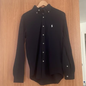 Mörkblå skjorta från Ralph Lauren - Snygg svart långärmad skjorta från Ralph Lauren med klassisk krage och vita knappar. Skjortan har en broderad logotyp på bröstet och är tillverkad i ett lättviktigt material. Perfekt för en stilren look.