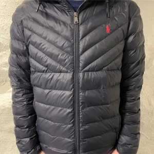 Svart dunjacka från Polo Ralph Lauren - Snygg svart dunjacka från Polo Ralph Lauren med huva och dragkedja. Jackan har ett diskret rött logotypbroderi på bröstet och är quiltad för extra värme. Perfekt för kyligare dagar.