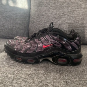 Nike Tn’s - Snygga Nike sneakers med ett unikt mönster i svart och grått. Skorna har en röd Nike-logga och svarta snören. Perfekta för dig som vill ha en stilren men ändå iögonfallande look. Såklart äkta