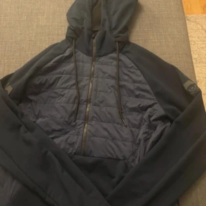  Sail Racing cardigan  - Snygg svart cardigan  från Sail Racing med dragkedja och huva. Framsidan har ett quiltat mönster och det finns en liten gul detalj på baksidan. Perfekt för en sportig look.