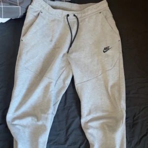 Grå mjukisbyxor från Nike - Snygga grå mjukisbyxor från Nike med svart snörning i midjan och praktiska fickor med dragkedja. Perfekta för en avslappnad stil eller träning. Nike-loggan är diskret placerad på benet.