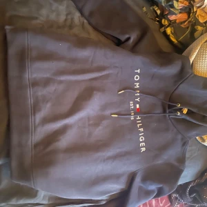 Svart hoodie från Tommy Hilfiger - Säljer en svart hoodie från Tommy Hilfiger med logga och texten 'EST. 1985' på framsidan. Har aldrig haft på mig den, säljer eftersom att jag köpte den som present till någon men de ville inte ha den. Nypris 1300