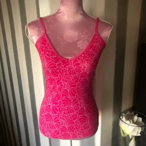 Rosa mönstrad linnetopp - Snygg rosa linnetopp med ett blommigt mönster. Toppen har smala axelband och en v-ringad design som ger en feminin touch. Perfekt för varma dagar eller som ett lager under en jacka. Med inbyggd bh