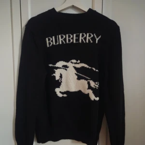 Svart stickad tröja från Burberry, S  - Snygg svart stickad tröja från Burberry med ett stort vitt logotyptryck och en riddare på framsidan. Storlek S. Oanvänd. Färg: Svart. 