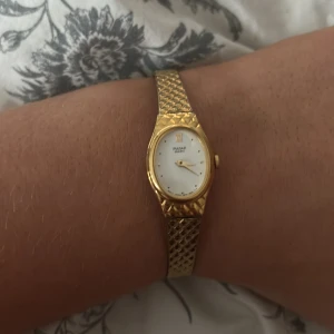 Guldfärgad klocka från Pulsar - Elegant guldfärgad klocka från Pulsar med en vit urtavla och guldfärgade visare. Armbandet har ett mönstrat design som ger en lyxig känsla. Perfekt som accessoar för att lyfta din stil.