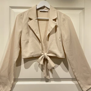Beige Topp - Säljer denna beiga toppen/koftan från NA-KD. Knytningen kan göras på flera olika sätt. Storlek 36. Säljes för 120kr. Fraktas på köparens bekostad