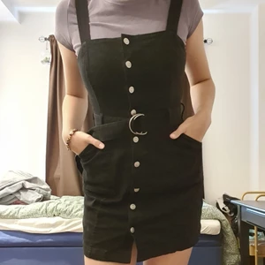 Svart overall-klänning - Overall-klänning av jeanstyg med silvriga knappar, fickor och matchande bälte till! Klädesplagget öppnas framifrån och längden på remmarna går att justeras. Snygg med t-shirt under eller långärmad tröja!