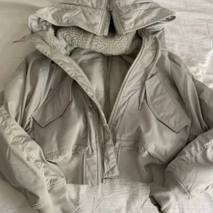 Vårjacka  - Ljus grå croppad bomberjacka från weekday. Silverdragkedjor på både armar, rygg och luva. Luvan är stor och täckt i vitt teddi tyg. Denna jacka passar våren men även hösten. Knappt använt och säljer nu pga ingen användning av den längre. 