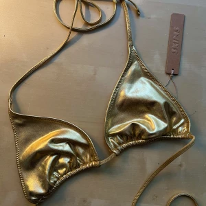 SKIMS bikini guld - SKIMS bikini i guld. Superfin, helt oanvänd men för liten för mig. Storlek M. Vanlig triangelbikini, knyt i ryggen och knyt i nacken. Inga ’pads’.  Vill inte lämna tillbaka då frakt tillbaka är så dyr. Kan mötas upp eller posta.  Tar ej emot returer. 