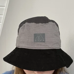 Bucket hat Buff - Svart och grå bucket hat från Buff. Köpt på Sellpy men den har aldrig kommit till användning. Spänne i bak som man kan reglera hur tight man vill att den ska sitta (se bild nr 3).
