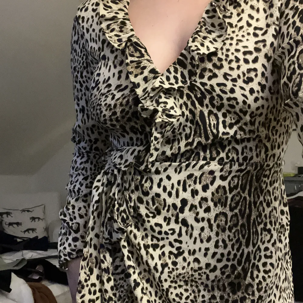 Leopardmönstrad klänning i omlott med volanger. Snöre i midjan och en hake vid bröstet så man inte behöver oroa sig över att visa för mycket. Använd typ en gång, slutar vid knäna.. Klänningar.