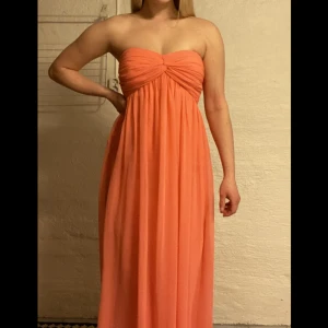 Balklänning  - Balklänning i korallfärg, det är svårt att göra färgen rättvisa på bild. Den sitter fast bra så man är inte rädd att den ska glida ned, går hela vägen ned till marken på mig som är 163. Använd en gång. 