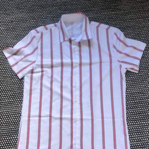 En väldigt fin kortärmad skjorta. Den är vit och röd. Den är i storlek M. Den är i väldigt bra skick då den aldrig har används!