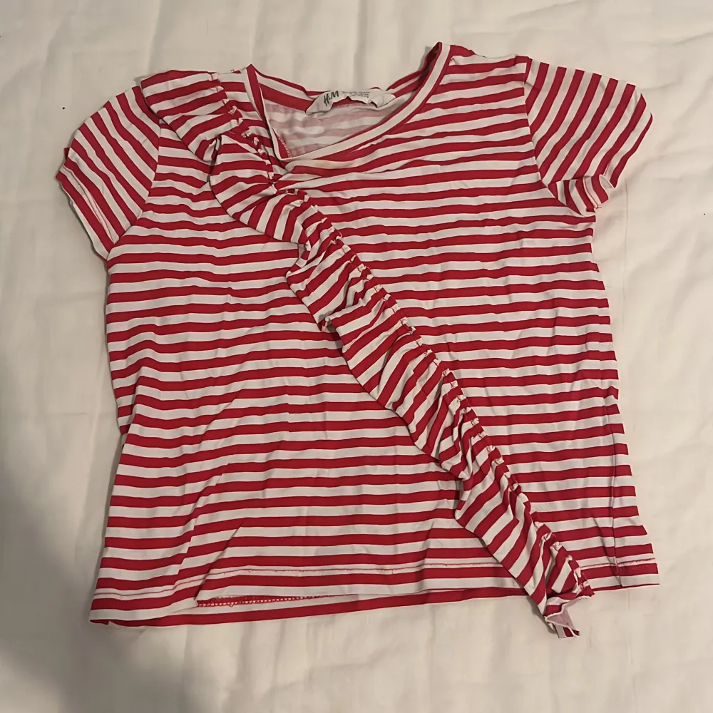 Säljer en t-shirt från hm. Den är röd vit och har en volang. Storlek 110-116 säljer pågrund av för liten. Den har ett litet hål vid bröstet. Kostar 15kr plus frakt. Står inte för postens slarv.💘. T-shirts.