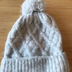 H&M grå stickad mössa - Grå stickad mössa med liten toffs från H&M. Passar fint för både höst å vinter samt håller värmen bra. 