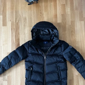 Gant dunjacka - Klassisk dunjacka Jacka vadderad med 90% dun och 10% fjäder. Den har en löstagbar huva, snedfickor i midjan, ribbstickade muddar och en broderad GANTt-sköld på vänster arm. Knappt använd, superfint skick.  