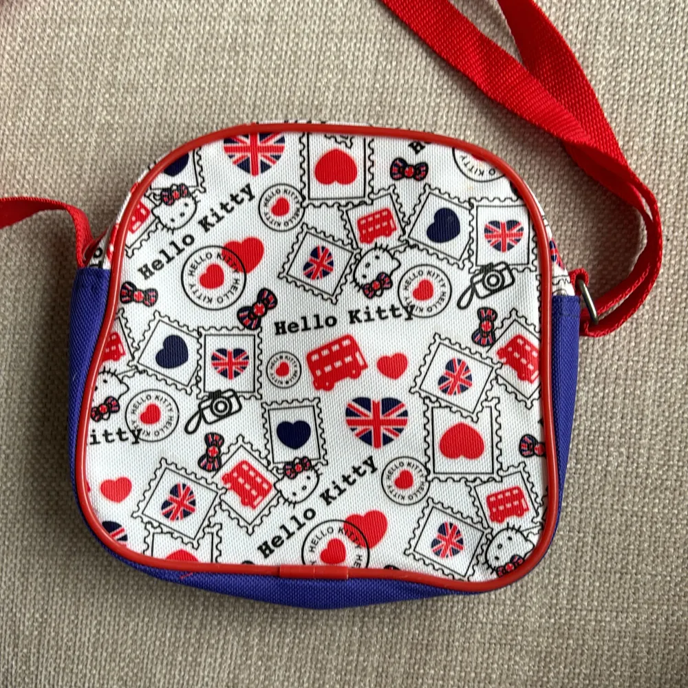 Söt liten väska med motiv av Hello Kitty Globetrotter collection -UK  Färger röd, vit och blå. Justerbar axelrem. Storlek ca 17x16 cm. Ett fack som stängs med dragkedja. . Väskor.