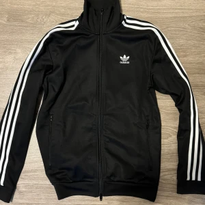 Svart Adidas kofta med i storlek S - Säljer en svart Adidas kofta i storlek S med klassiska vita ränder på ärmarna och Adidas-logga på bröstet. Koftan är i mycket bra skick och är perfekt för träning. Bekväm passform.