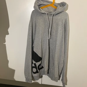 Adidas Hoodie - En Adidas hoodie som är ljus grå och har ett stort svart assists märke längs höger midja.  Köpt på stadium för ungefär 3 eller 4 år sen!  Vid snabb affär får du den för 60kr😊
