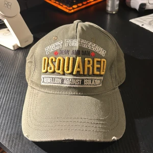 Grön keps från Dsquared2 - En grön keps från Dsquared2 med texten 'FIGHT FOR FREEDOM' och 'DSQUARED' broderad i vitt och gult på framsidan. Kepsen har även texten 'KILLERS ON THE LOOSE' broderad på baksidan. Den har rivningar i framsidan på kepsen som design. Pris kan diskuteras!