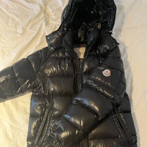 Svart dunjacka från Moncler - En svart Maya dunjacka från Moncler med huva och dragkedja. Jackan har en glansig yta och är quiltad för extra värme. Perfekt för kalla vinterdagar. kvitto och prislapp finns även. och sedan en påse om man vill ha med den, men de är ganska sliten. en liten skavank och de är att serieteckningen lossnat lite på undersidan men inget man märker av 